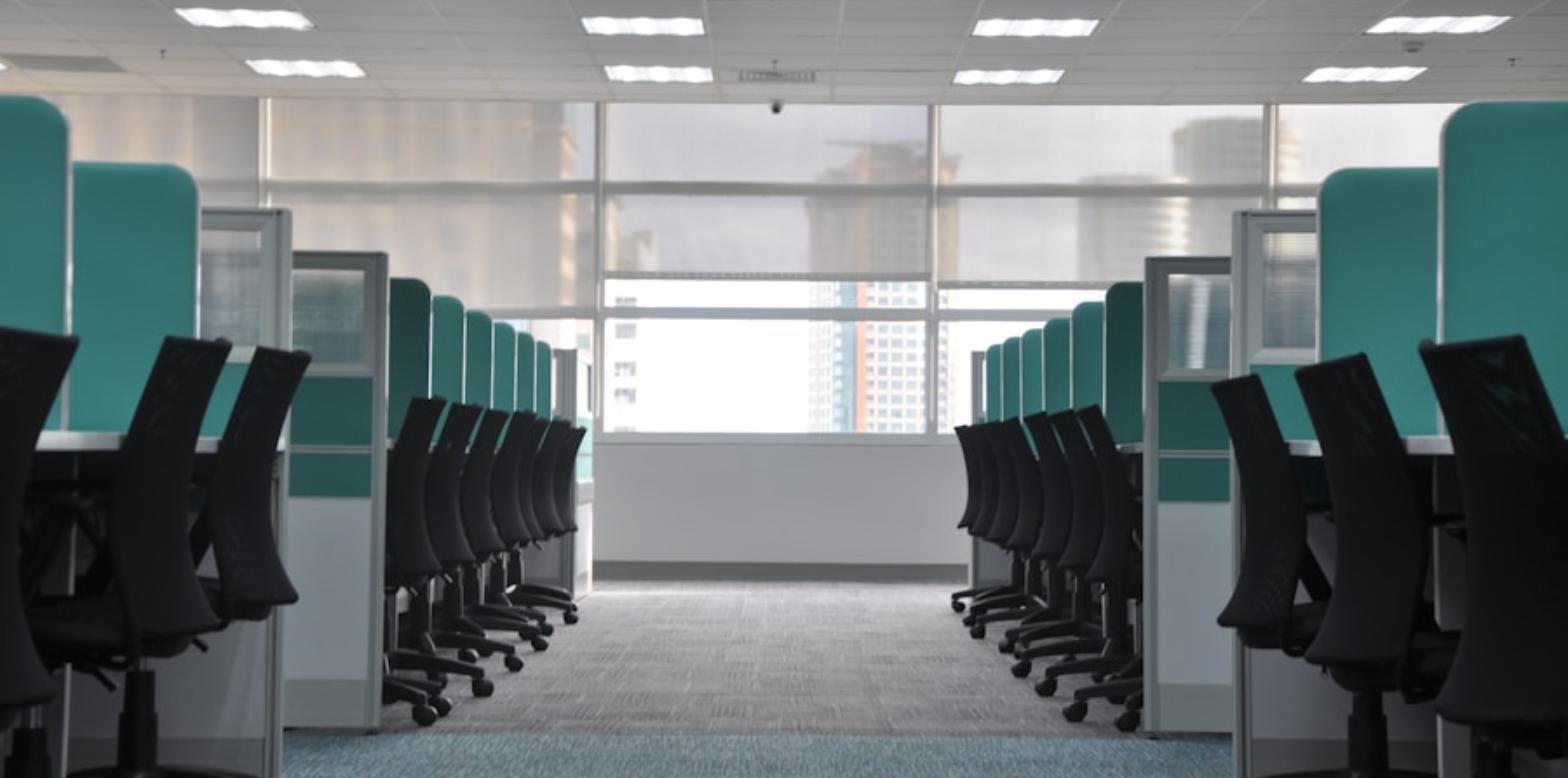 Fila de escritorios y sillas de oficina vacíos en cubículos, con un puesto de trabajo sin ocupar