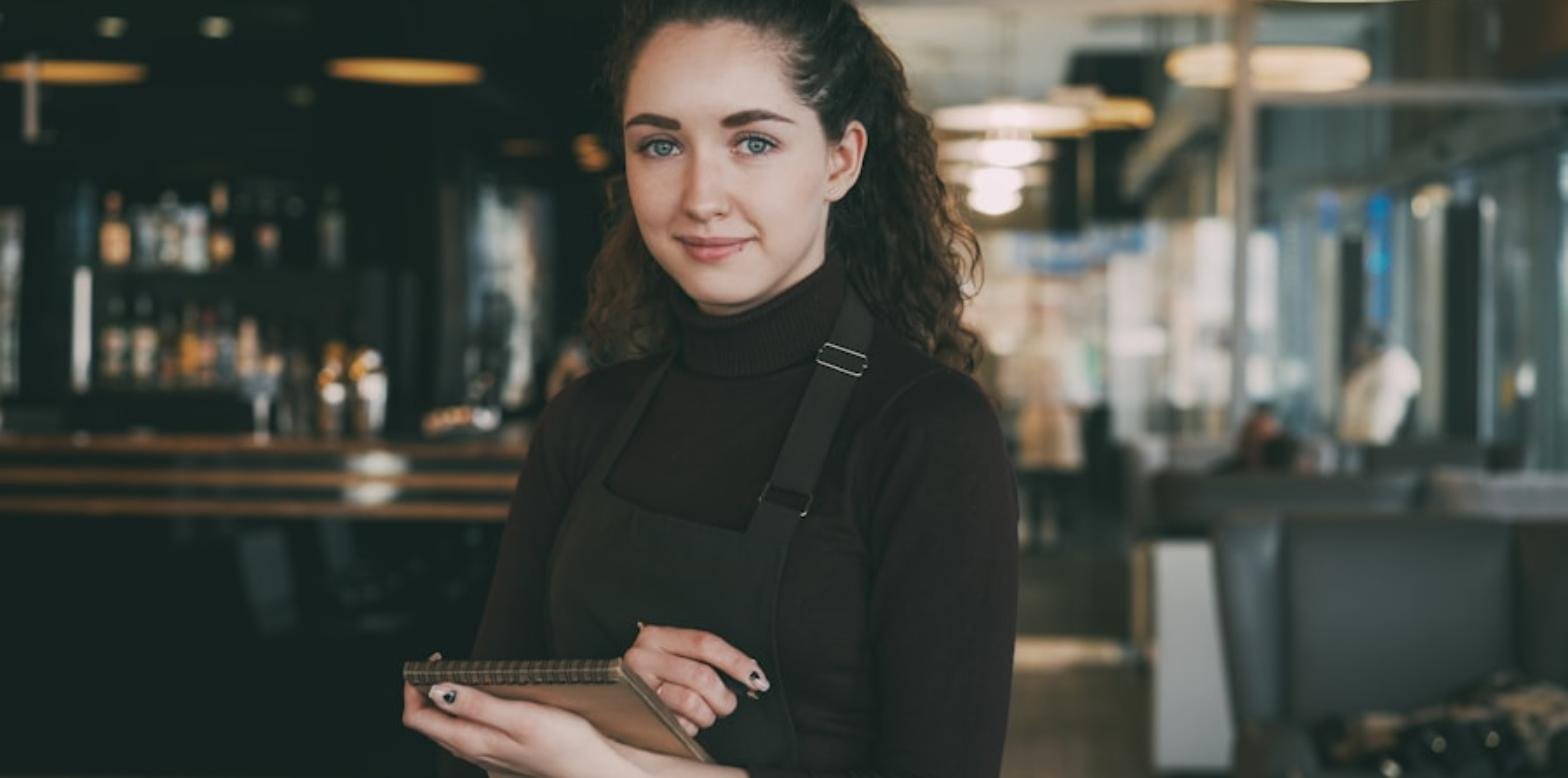 Una trabajadora de hostelería con delantal sostiene una libreta en un restaurante