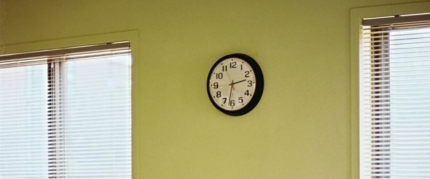 Un reloj de pared en una sala vacía con ventanas y persianas, simbolizando la larga pausa de la jornada partida