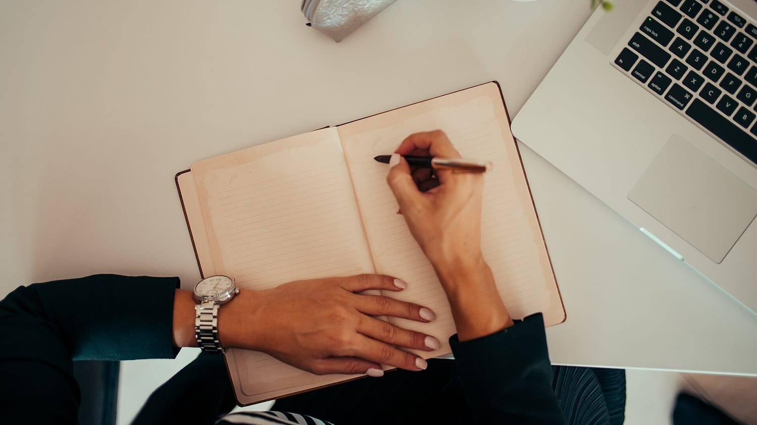 Manos de una persona escribiendo en una libreta junto a un ordenador portátil en una mesa de trabajo